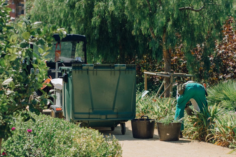 A gardener wearing teal overalls is crouched down amid lush garden beds filled with green plants and tall, spiky foliage, engaged in planting or trimming. To the left, a large green waste bin is posit